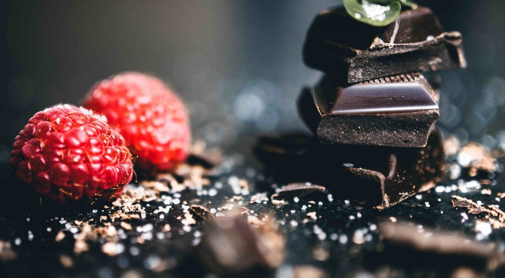 Bild: Schokoladenkurs - 4 Stücken dunkle Vollwertschokolade aufeinander gelegt, mit 2 Himbeeren daneben.