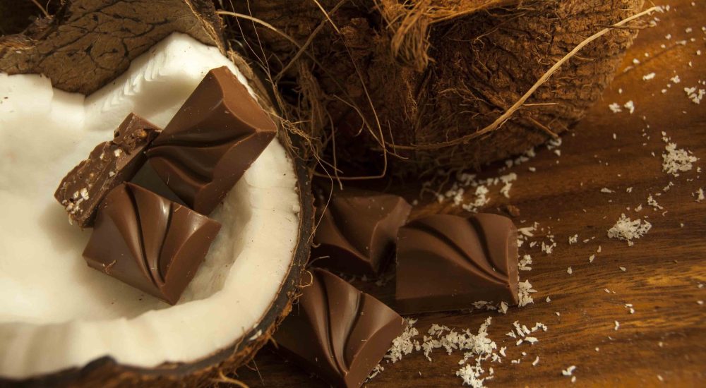 Bild: Schokoladenkurs - 2 Kokospralinen liegen auf dem weissen Fruchtfleisch einer offenen Kokosnuss auf einem braunen Tisch.